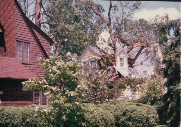 A two-story house with tornado damage to its roof and dormer, surrounded by downed trees