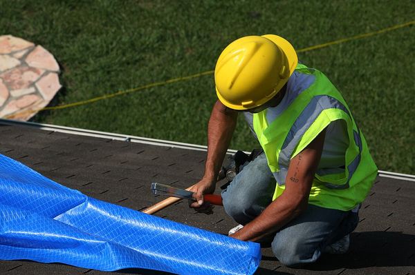 A worker in a hard hat nails a blue tarp to a roof with furring strips