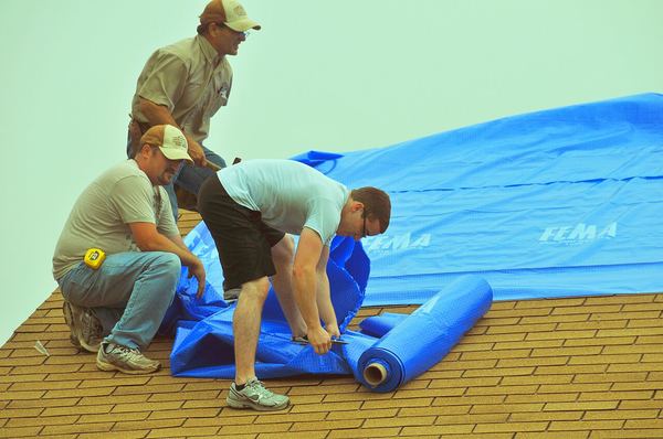 Three men unroll a large blue tarp over a damaged shingle roof